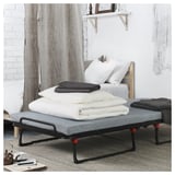 Modern bedroom with white bed, grey fabric guest bed, red feet, folded blankets on top. White curtains, wooden shelf with mirror.