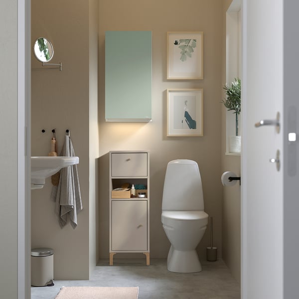 Small, beige NORSÄLVEN cabinet fits tight spaces. Compact with drawer and shelves for bathroom storage. Positioned between sink and toilet.