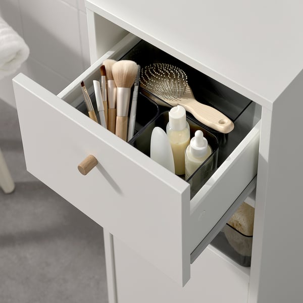 Open bathroom cabinet showing makeup brushes, hair tools, and bottles. Slender design offers organized storage with a drawer and shelves.