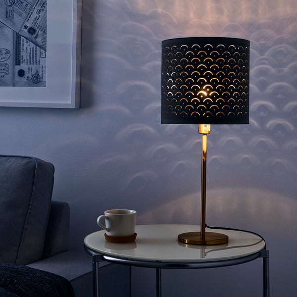 A modern lamp with a perforated shade casts intricate patterns on the wall. It sits on a round side table, illuminating a cozy living space.