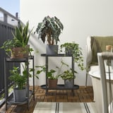 Black OLIVBLAD plant stand holds multiple pots, showcasing tall plants. Its positioned between a railing and a white chair, perfect for small spaces.