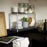 Kitchen counter with OMAR silver storage rack holding jars, bowls, and a plant. Wooden cutting board and bottles nearby.