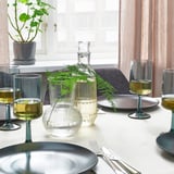 Table set with OMBONAD blue plates, clear textured glass vases holding greenery, and wine glasses on white tablecloth near window.