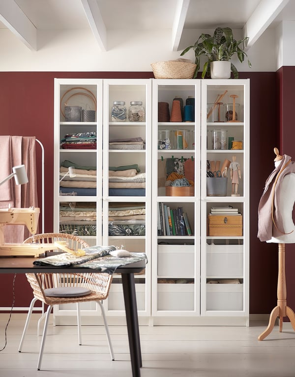 White OXBERG bookcase with glass doors, holding various items, set against a red wall in a cozy workspace with a table and chair.
