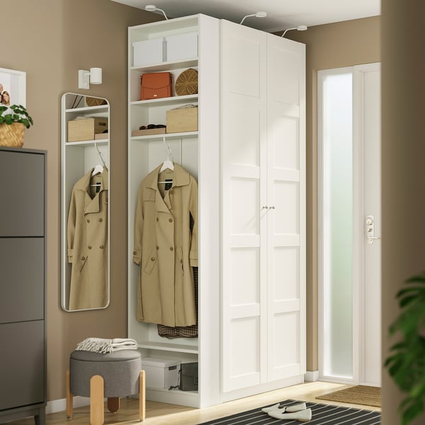 A white PAX wardrobe end-unit beside a door. It has narrow and tall, with garments hanging and accessories on shelves.