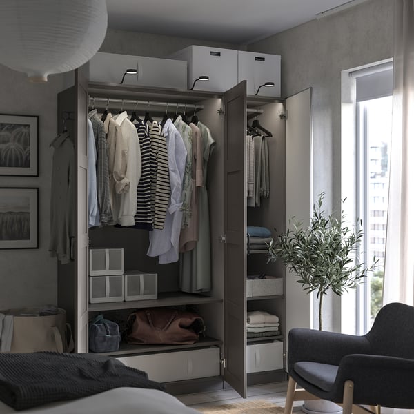 Modern bedroom an open wooden PAX wardrobe with clothes, shelves, and drawers; a black chair and a plant beside a window.