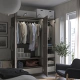 Modern bedroom an open wooden PAX wardrobe with clothes, shelves, and drawers; a black chair and a plant beside a window.