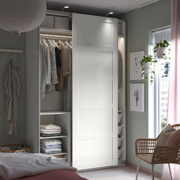 Modern bedroom with open PAX wardrobe, neatly folded clothes, hanging garments, organized storage, plants, and window decor.