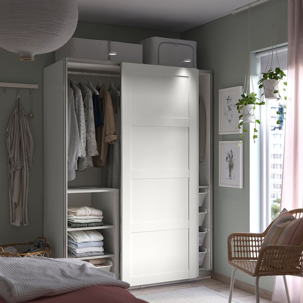 Modern bedroom with white IKEA PAX wardrobe, wicker chair, plant, and artwork.