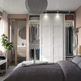 A modern, minimalist bedroom featuring a white PAX wardrobe with sliding doors, housing clothes and shelves.