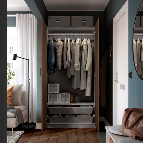 Open wooden wardrobe displaying clothes on hangers, shelves with boxes and drawers, set against dark walls.