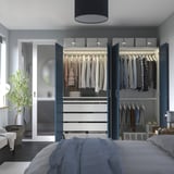 A modern bedroom with a PAX wardrobe, open to show organized clothes, and a cozy bed with a lamp and plant.
