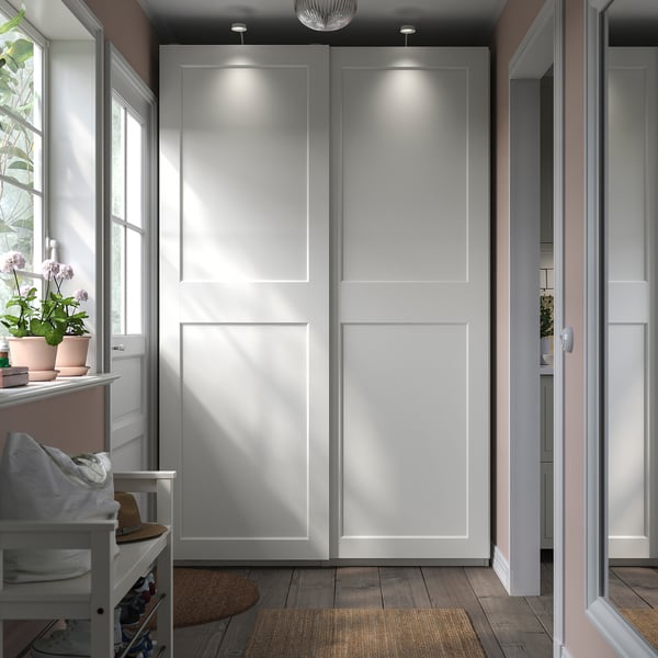 Bright, modern white wardrobe with sliding doors, lit panels, and a cozy corner with a chair and plants.