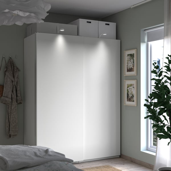 A modern bedroom a white PAX wardrobe with sliding doors and storage boxes. Greenery adds freshness and natural light brightens the space.
