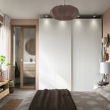 Modern bedroom with white PAX wardrobe, sliding mirrored doors, brown pendant light, brown bedding, nightstand with lamp, and a plant.