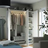 A modern bedroom with a white PAX wardrobe featuring clothes on hangers, folded items on shelves, and mesh baskets for storage.