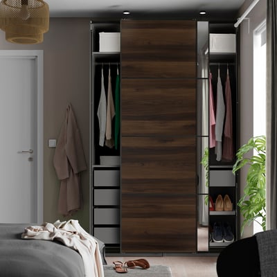 PAX wardrobe in dark wood next to bed, sliding doors reveal shelves and hanging clothes.