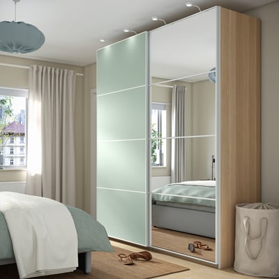 Modern bedroom with light wooden PAX wardrobe and mirrored doors reflecting window and bed. Soft lighting, beige curtains, and minimalist decor.