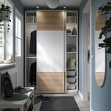 Modern closet with white and wood texture sliding doors, hanging clothes, shelves, baskets, and a round mirror.