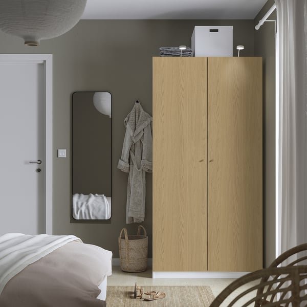 A minimalist bedroom with a tall light wooden wardrobe, mirror, and wicker basket. A bed with white linens is partly visible.