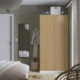 A minimalist bedroom with a tall light wooden wardrobe, mirror, and wicker basket. A bed with white linens is partly visible.