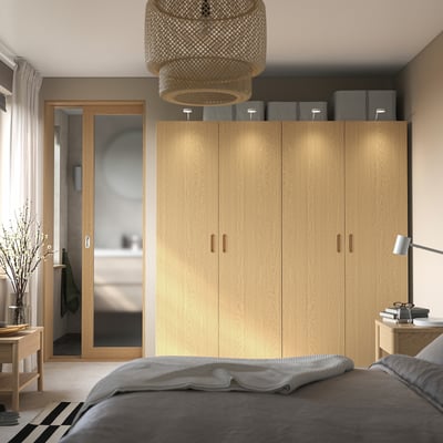 Modern bedroom with light wooden PAX wardrobe, grey linen, and hanging pendant light. Wardrobe stands tall beside bed, with grey storage boxes on top.