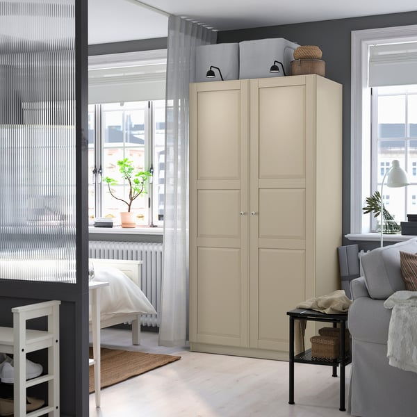 Bright bedroom with large beige PAX wardrobe, white furniture, grey couch, and potted plant, with large windows.