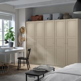 A beige PAX wardrobe in a room, surrounded by a black desk, chair, and coffee table. Near windows, with decor and a bed in front.