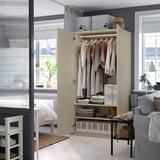 Open beige PAX wardrobe with clothes; hinged doors closing. Small white bed, sheer curtains nearby.