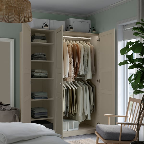 A beige PAX wardrobe holds clothes on hangers and folded on shelves. Integrated lighting illuminates the interior.