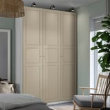 Large beige PAX wardrobe with soft-close hinges, positioned near a window with green plant, chair, and bed.