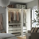 A modern, open wardrobe with clothes hangers, drawers, and shelves. It integrated lighting and is set against a neutral wall with a sofa nearby.