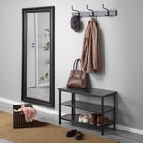 A hallway setup with a wall-mounted coat rack holding a hat and coat, a shoe bench with shoes and a handbag, and a large mirror beside it.