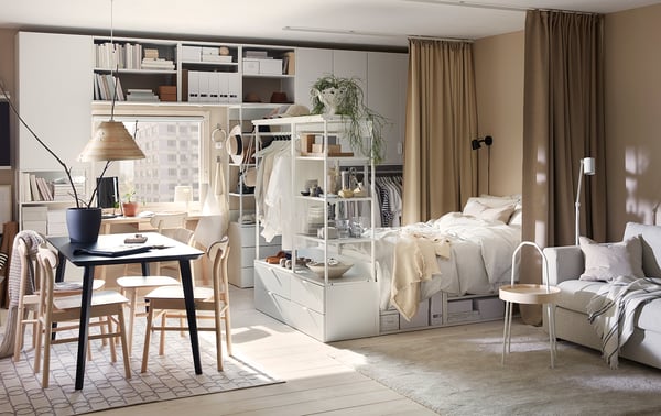 Modern studio apartment featuring PLATSA bed in white, combining sleep and storage, with beige curtains, wooden dining set, and tasteful decor.