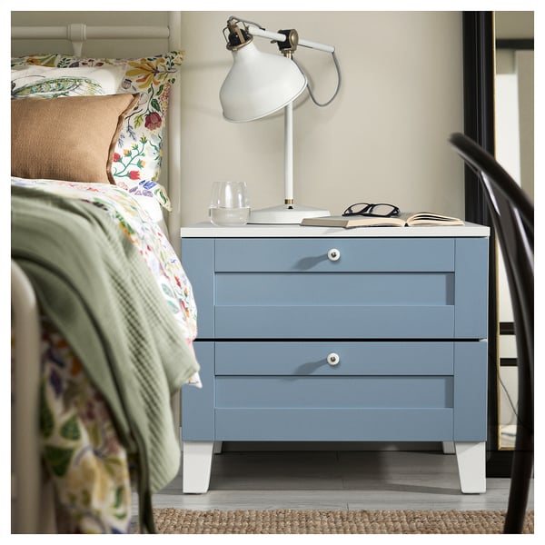 Cozy bedroom with blue PLATSA nightstand, lamp, books, floral pillows, green blanket.