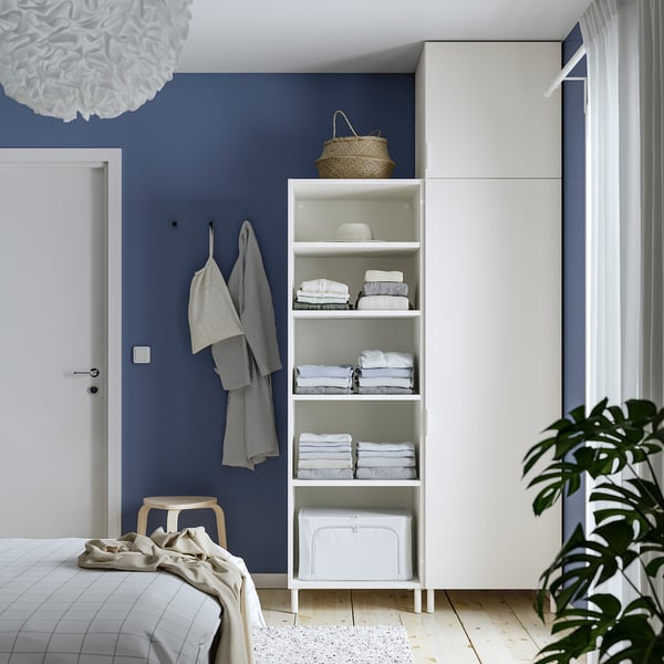 Modern bedroom with white PLATSA storage, towels, seagrass basket, wicker towel, and green plants against blue wall.