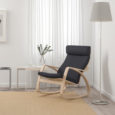 Modern living room with dark blue POÄNG rocking chair, light wooden frame, high back, and grey lamp. Natural light, beige rug, and side table.