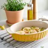 Yellow stoneware baking dish with lemons and herbs, potted plant, oil bottle. Round, textured, with handles.
