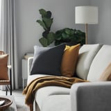 Modern living room with white couch, gray, and mustard yellow pillows, a cozy brown blanket, and a large green plant beside a lamp.