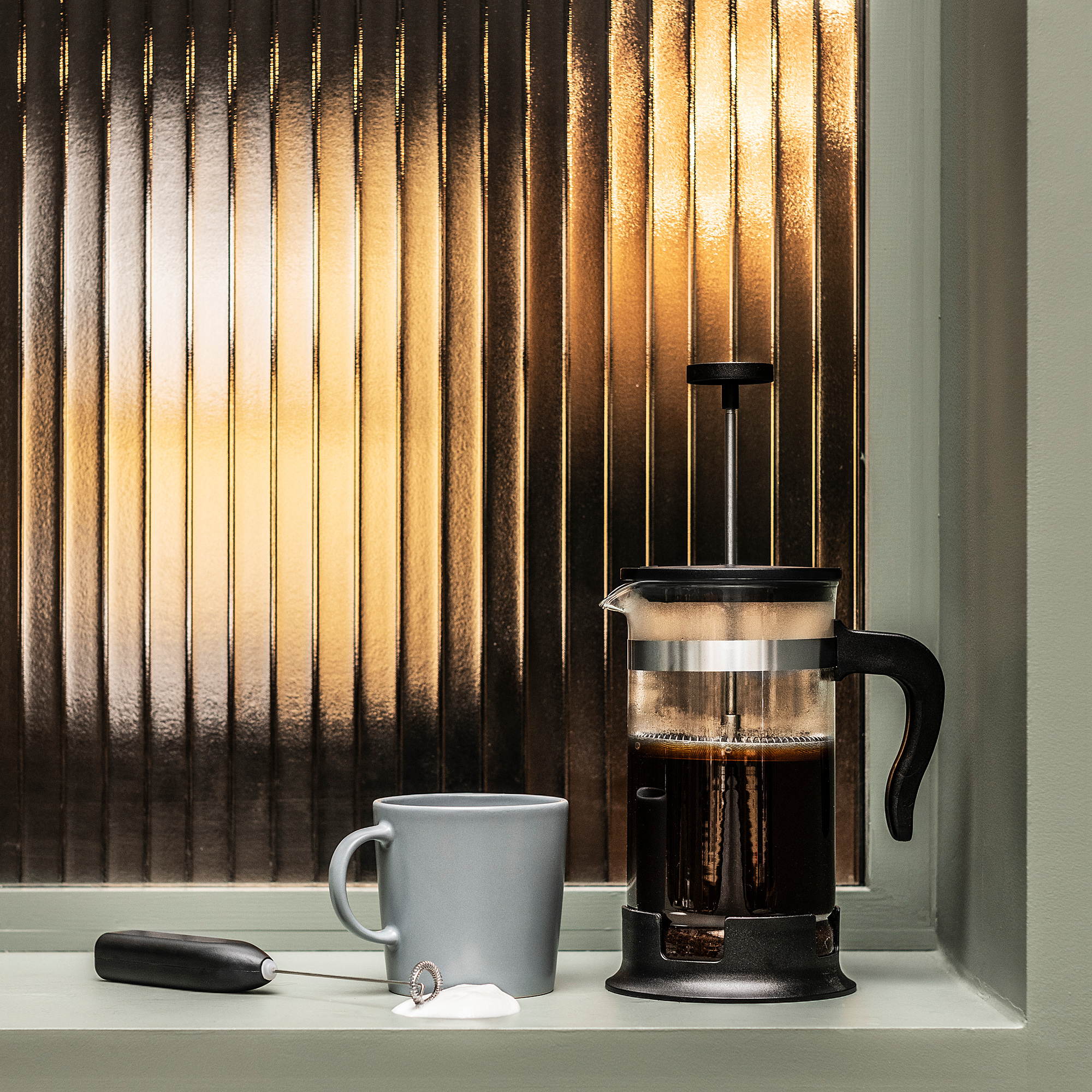 Black french press coffee maker on window sill with gray mug, frothing wand, and velvet bag near window with vertical blinds.