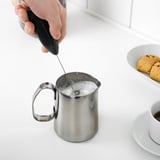 Hand holds black frother making foam in metal pitcher on white counter, beside coffee and cookies.