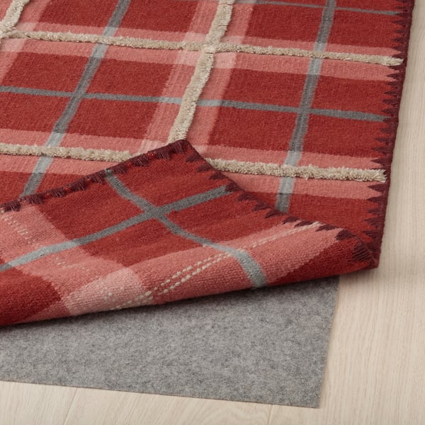 Red checkered wool rug folded on light wooden floor with gray rug pad underneath.