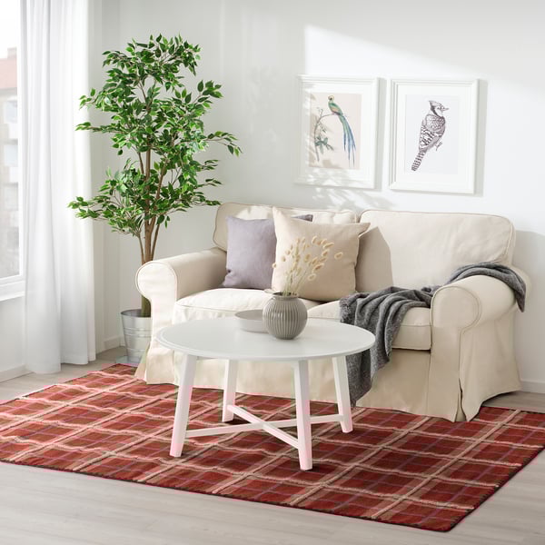 Large red wool rug with tartan pattern under beige sofa, white coffee table, and potted plant. Brightens cozy living space.
