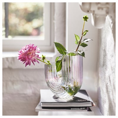 Clear glass vase RÄFFELBJÖRK with grooved design holding pink flower by window on books.
