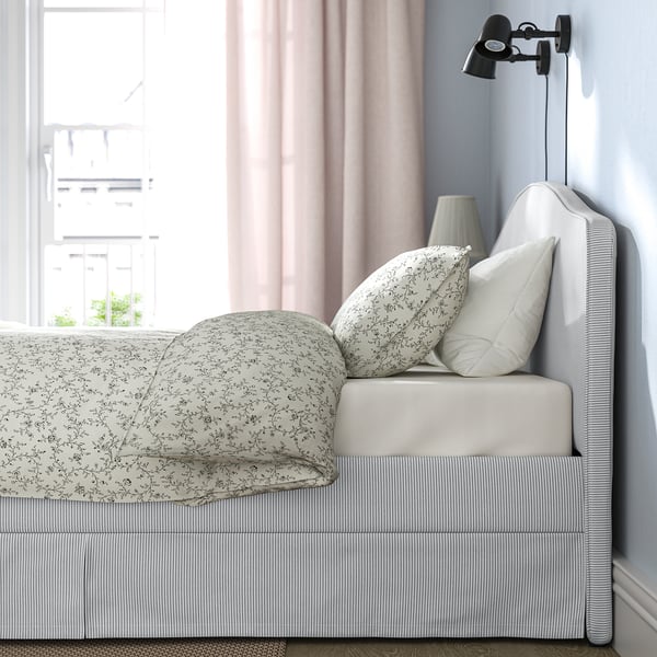 A cozy bed with a curved headboard, striped bed skirt, and floral pillows. The headboard has a wall-mounted light, offering functional reading light.
