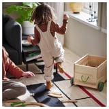 A child playing with wooden toys and an adult on a rug. A wooden cart with green wheels is nearby. The rug is beige and black with red edges.