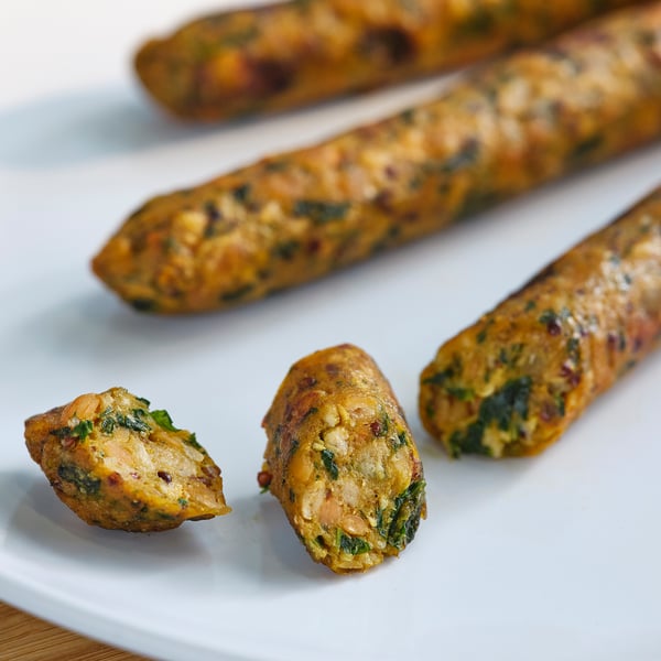 Three brown, cylindrical vegetable sausages on a white plate, with one cut in half, revealing green flecks.