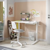 A white sit/stand desk with a green shelf and lamp, paired with a white chair, set against a peach wall.