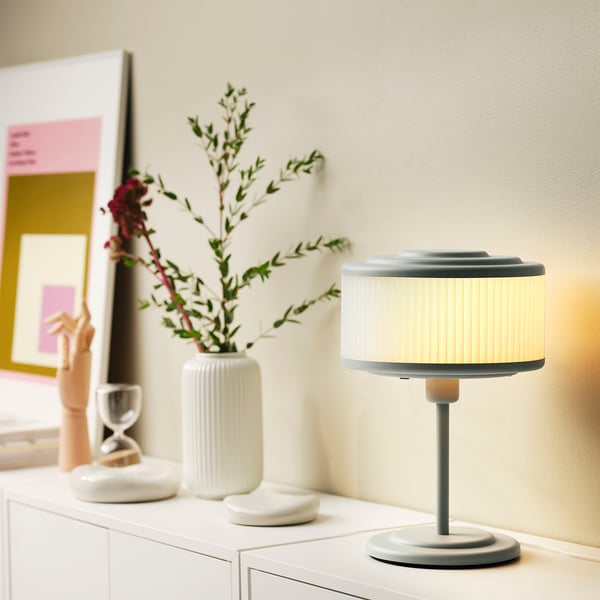 Modern minimalist desk setup. Small grey REMTÅNG lamp, white vase with floral, hourglass on shelf. Lighted lamp highlights warm decor.