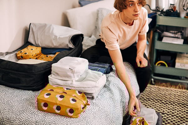 A young person packs clothes into yellow and black patterned RENSARE packing cubes on a bed. The cubes are neat and organized, ready for travel.